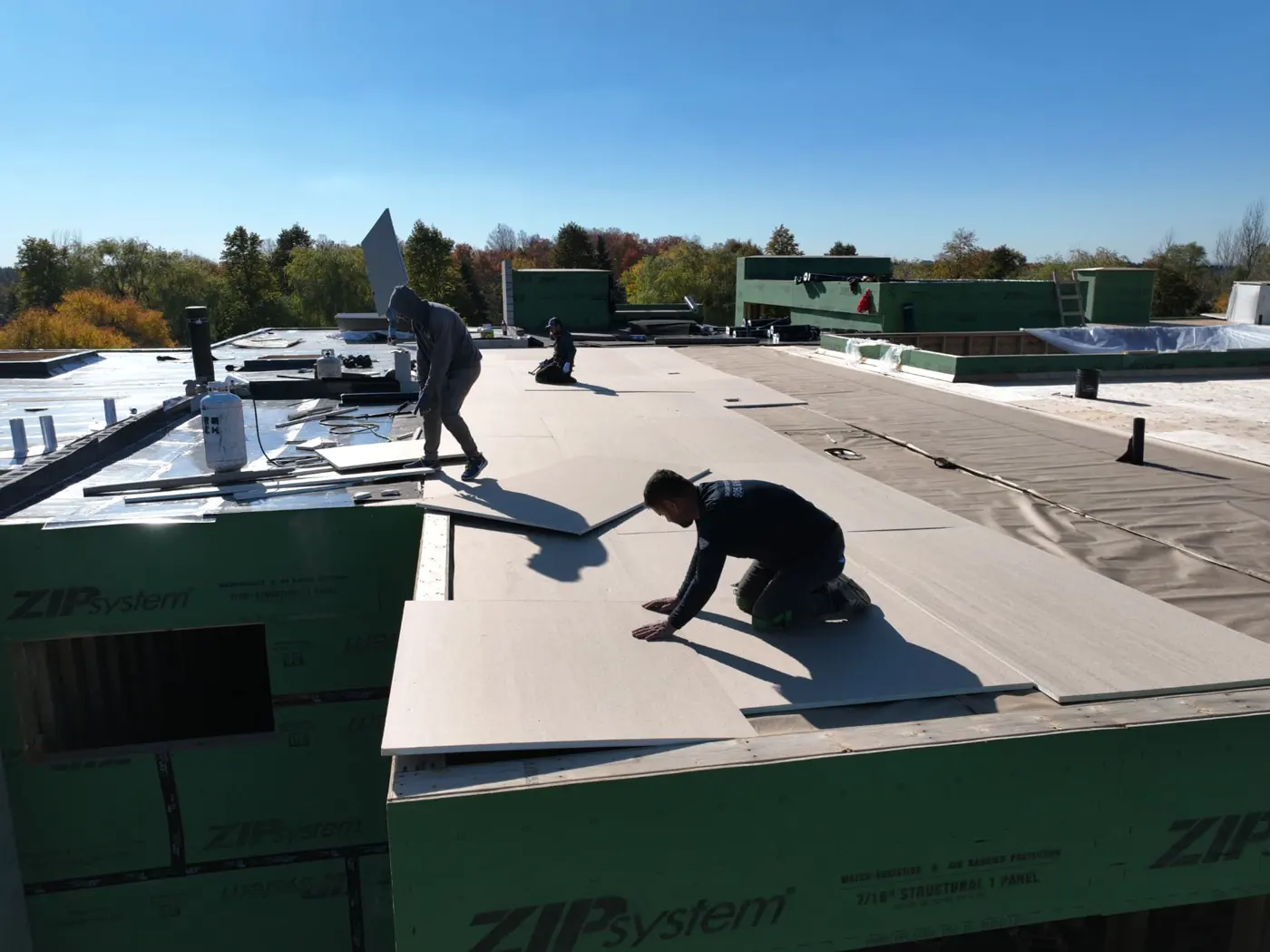 Close drone view of a roofer kneeling to align a rigid board at the edge of a Brampton flat roof while another crew member carries a board