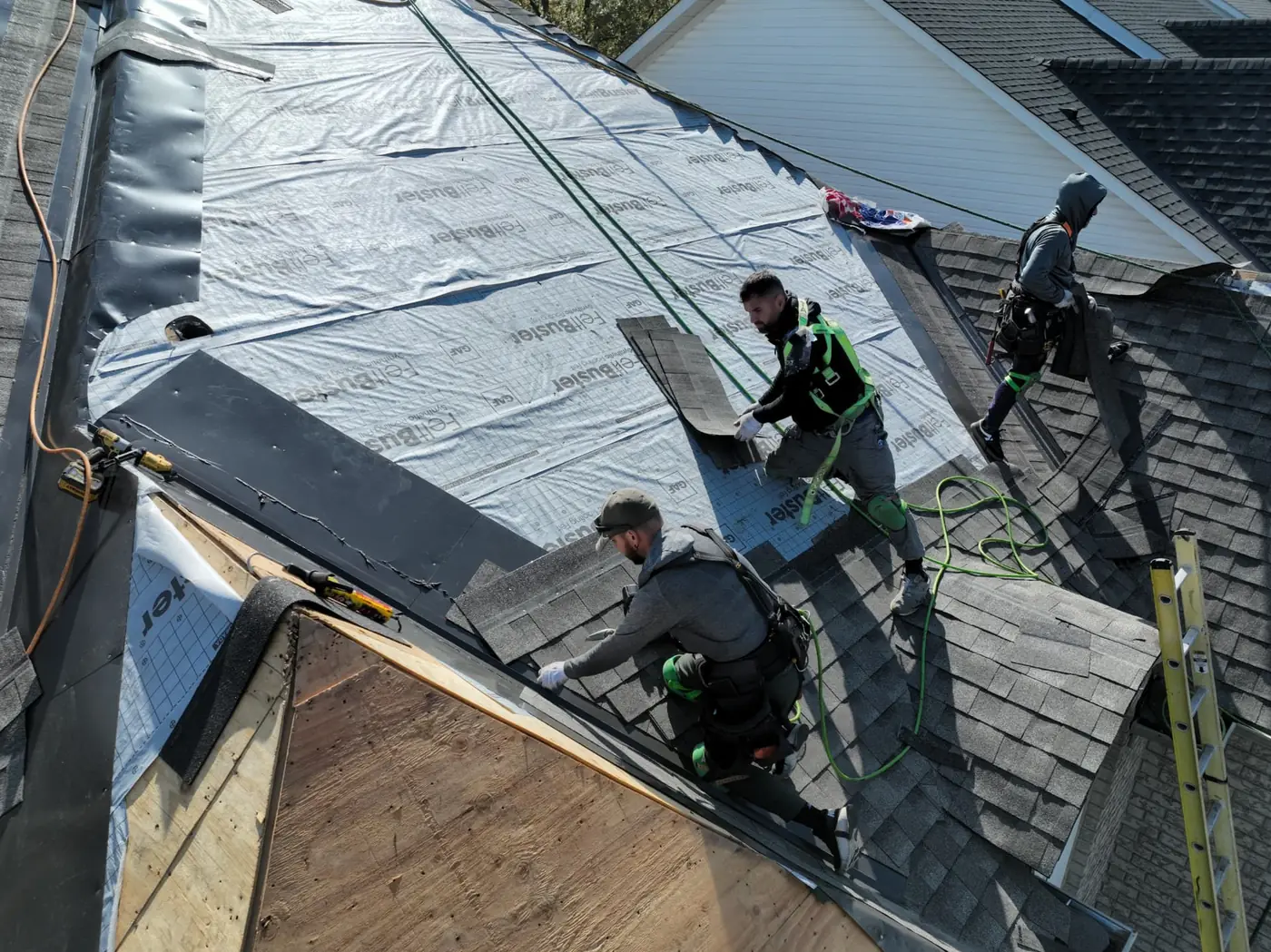 Three roofers laying dark shingle courses along a valley on an underlayment-covered Oakville roof with an exposed plywood section below