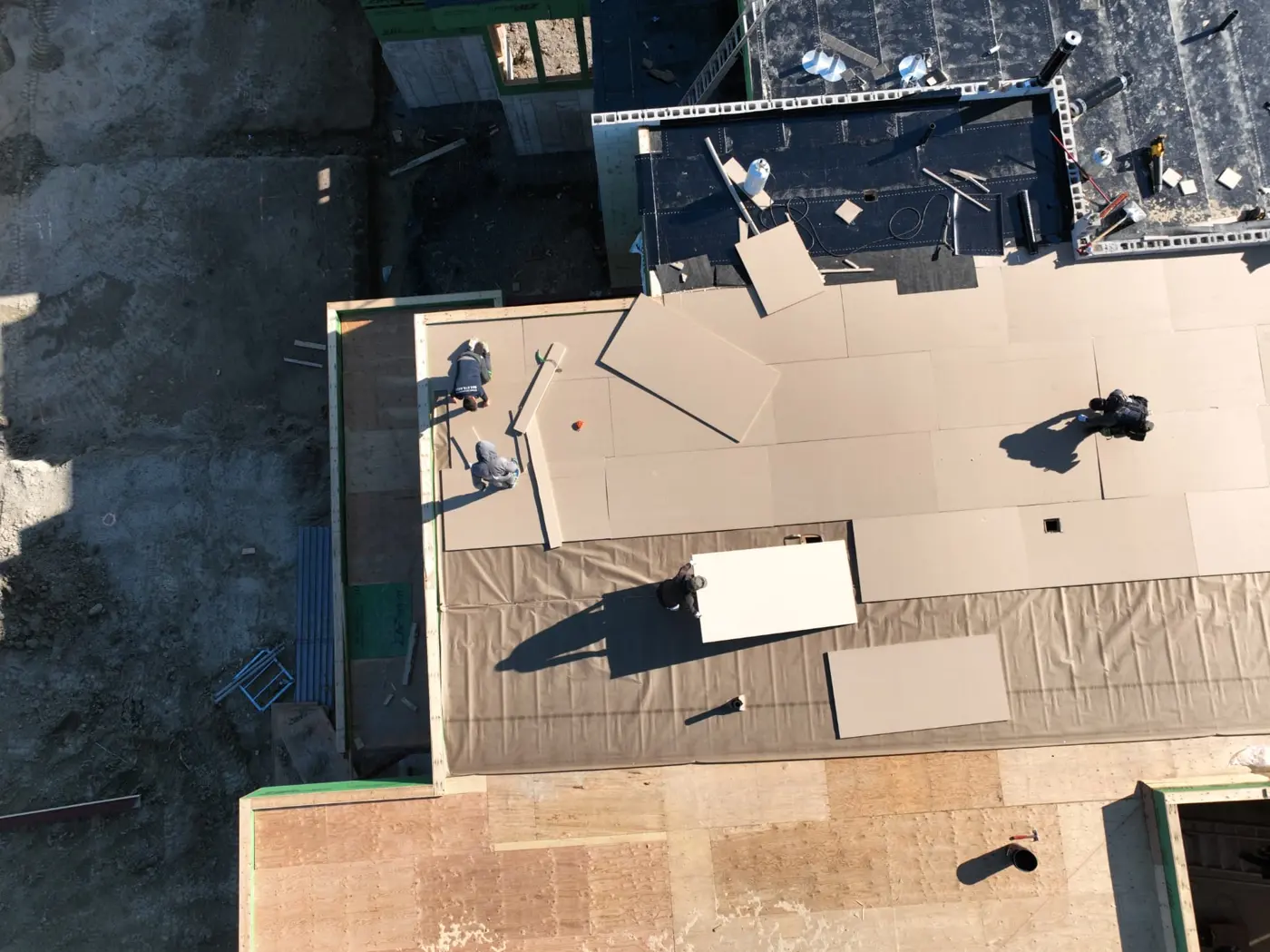 Top-down drone view of a Brampton roofing crew laying rigid boards over a flat roof, with loose boards, exposed decking and adjacent black membrane