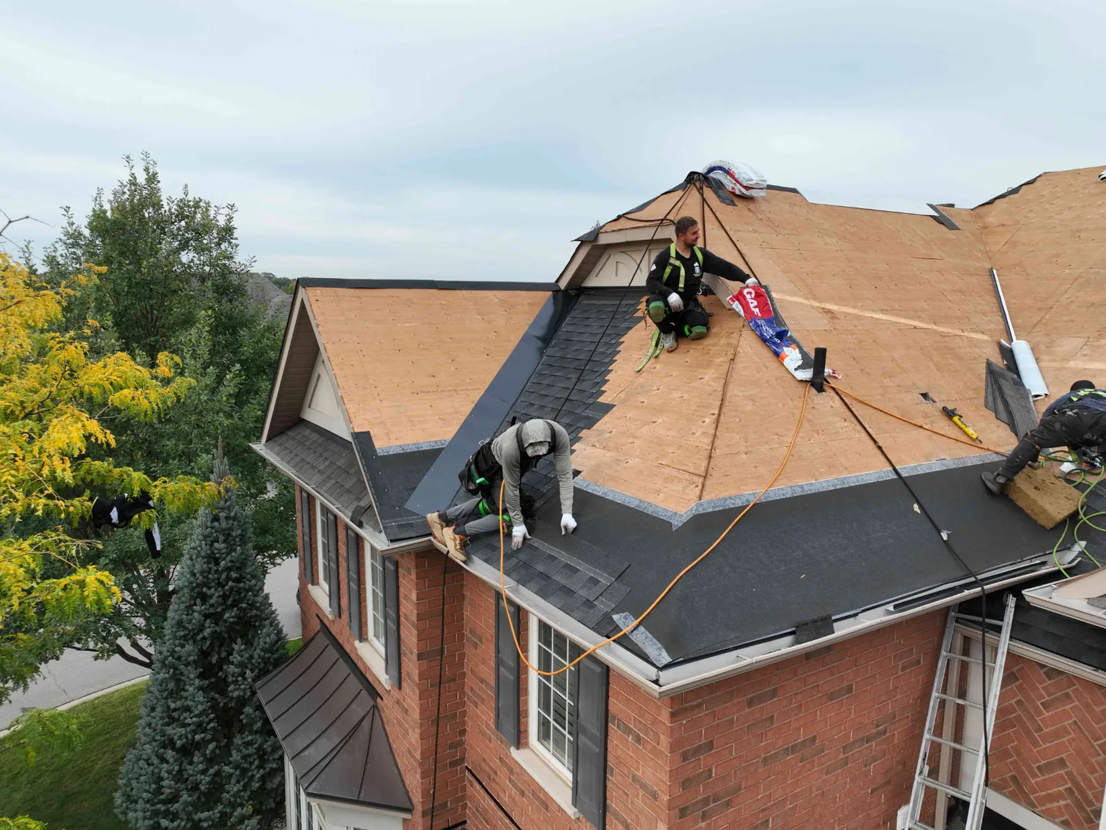 Drone view of roofers installing dark architectural shingles over black underlayment on a two-storey brick Oakville home