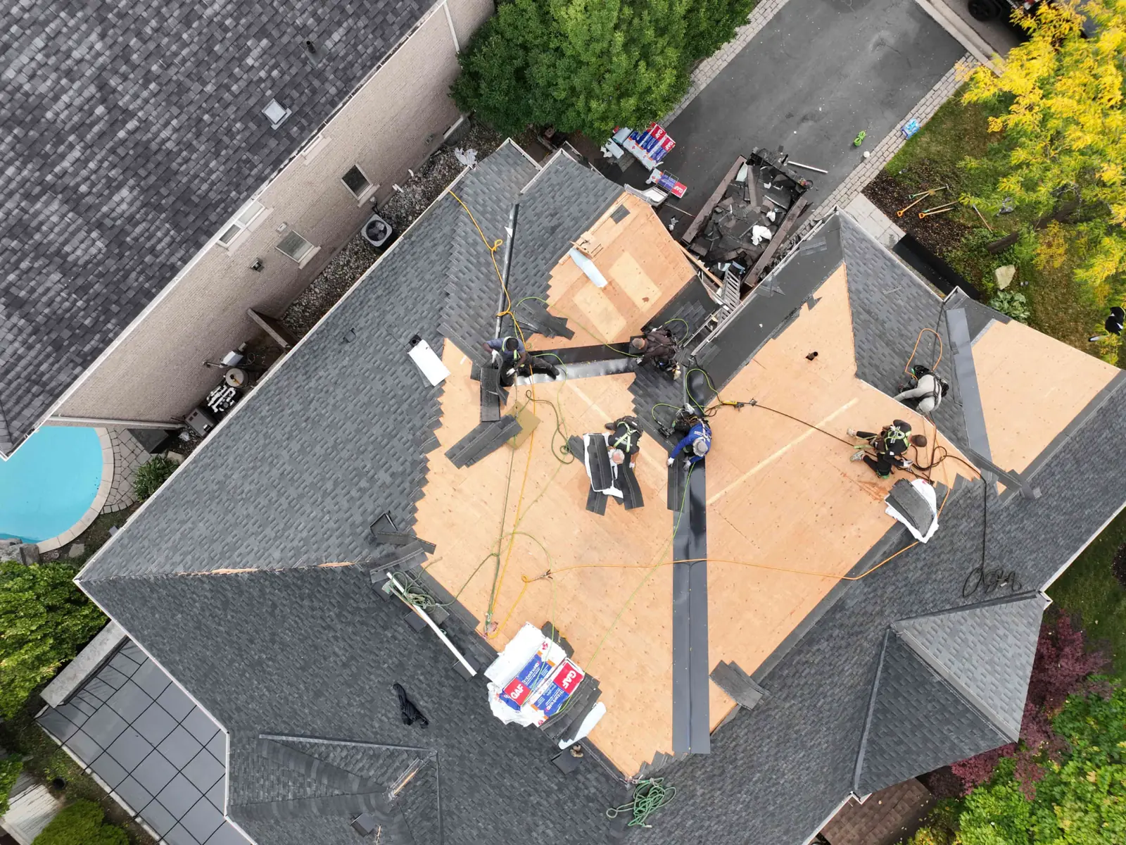 Top-down drone view of an Oakville roof replacement with exposed plywood decking, black underlayment strips, staged shingle bundles and a trailer of torn-off material